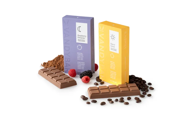 Two VandyBar boxes, lavender Raspberry Dreamin’ and yellow Rise n’ Grind, with unwrapped milk chocolate bars, raspberries, cocoa powder and espresso beans